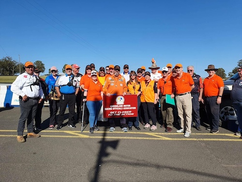 A group shot of a number of members at a motorsport event. They are holding a sign with the club logo, name, and tagline "Get closer to the action"